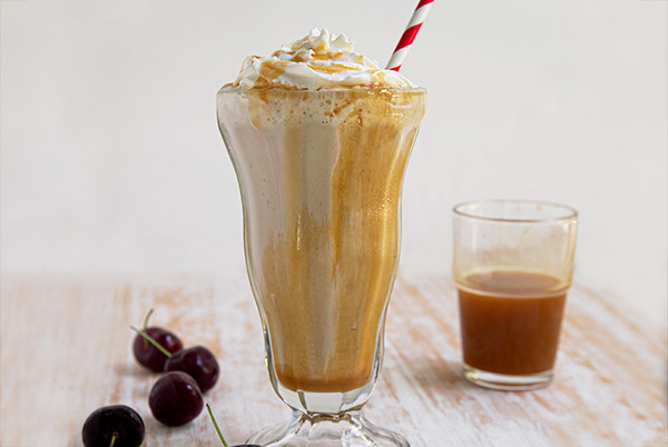 SunButter and Caramel Milkshake with a white and red striped straw on a beige and white table with a cup of caramel sauce and cherries in the background