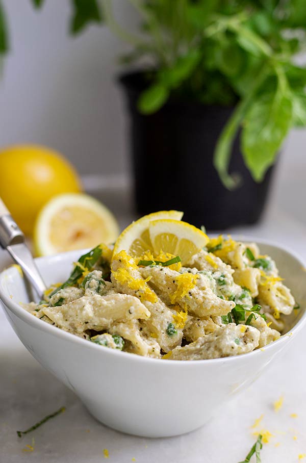 Lemon Ricotta Pesto Pasta in a white bowl with fresh lemons and a potted basil plant in the background