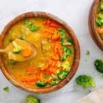 Overhead view of Plant-Based Broccoli 'Cheese' Soup in a brown bowl with a spoon lifting soup out of the bowl