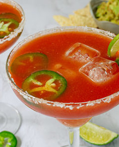 Closeup of Spicy Salsa Margaritas with salt on the rim of the glasses and jalapeno slices and lime garnishes with chips and guacamole in the background
