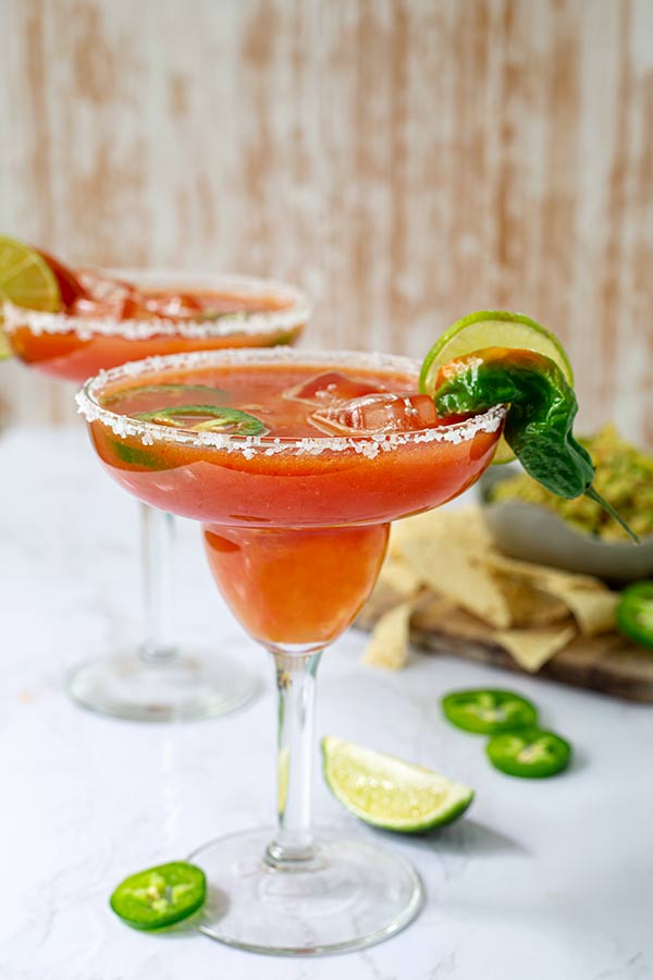 Two Spicy Salsa Margaritas garnishes with jalapeno slices and lime on a white table with wood wall background and chips and guacamole in the back