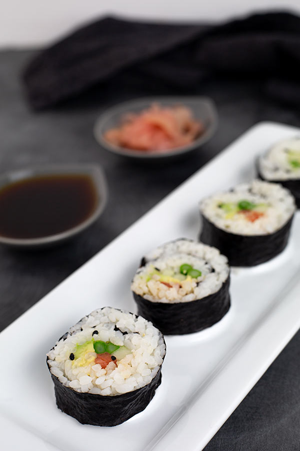 Closeup of Vegetarian Sushi on a white rectangular platter on a dark gray table with a ramekin of soy sauce and pickled ginger in the background