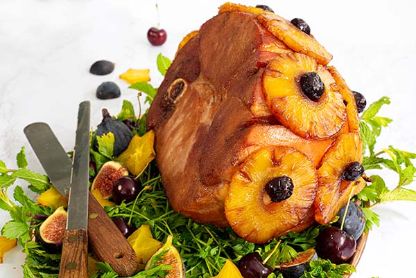 Close-up of Angela's Ham dotted with pineapple and cherries on a bed of greens with figs and cherries surrounding the ham