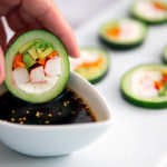 Closeup of a hand dipping Cucumber Sushi into soy sauce in a white ramekin