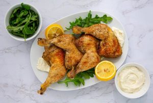 OVerhead view of Garlic-Lemon Chicken Legs on a white oval platter garnished with arugula and lemon with sauce on the side on a white marble countertop