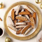 Overhead view of Gingerbread Biscotti on a white and gold plate with gold ball ornaments and cinnamon sticks and a cup of coffee surrounding the plate
