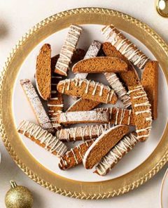 Overhead view of Gingerbread Biscotti on a white and gold plate with gold ball ornaments and cinnamon sticks and a cup of coffee surrounding the plate