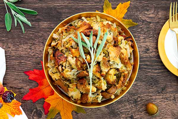 Overhead view of Mom's Stuffing in a gold bowl on a dark wood table with orange maple leaves under the bowl and sage garnish