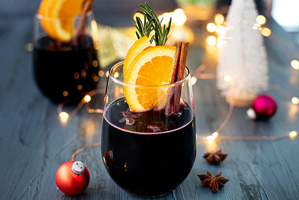 Mulled Wine in two stemless wine glasses with cinnamon sticks and orange slices inside for garnish on a dark gray table with holiday decorations around it