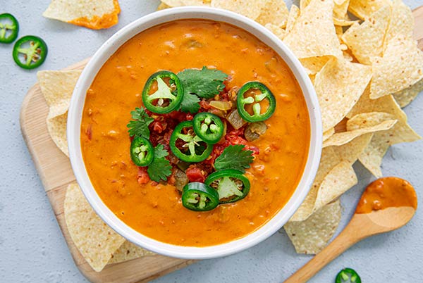 Overhead view of Plant-Based Chorizo Queso in a white bowl topped with tomato and jalapeno slices with corn tortilla chips surrounding the bowl set on top of a light wood cutting board