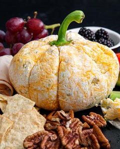 Cheese ball in the shape of a pumpkin with grapes and veggies and nuts around it against a black background
