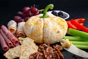 Cheese ball in the shape of a pumpkin with grapes and veggies and nuts around it against a black background