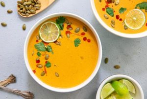 Overhead view of Thai Pumpkin Soup in white bowls garnished with lime slices and pumpkin seeds on a gray background