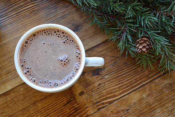 Overhead view of Bare Life Hot Chocolate in a white mug on a wood table with Christmas tree branches on the side