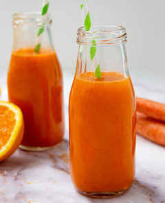 Carrot Apple Ginger Juice in two glass bottles with green and white striped straws in them with an orange sliced in half and two carrots next to the juice bottles on a white marble countertop