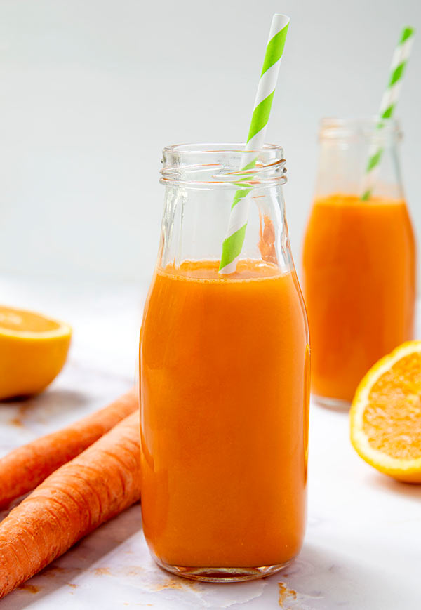 Closeup of two bottles of Carrot Apple Ginger Juice with green and white striped straws with fresh carrots and orange halves next to the bottles against a light gray background