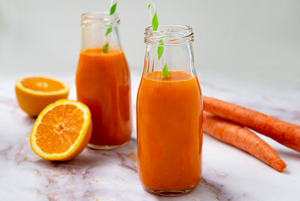 Carrot Apple Ginger Juice in two glass bottles with green and white striped straws in them with an orange sliced in half and two carrots next to the juice bottles on a white marble countertop