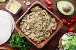 Overhead view of Fast or Slow Green Chili Shredded Chicken in a square wooden bowl on a wooden table surrounded by tomatoes and avocado and cilantro