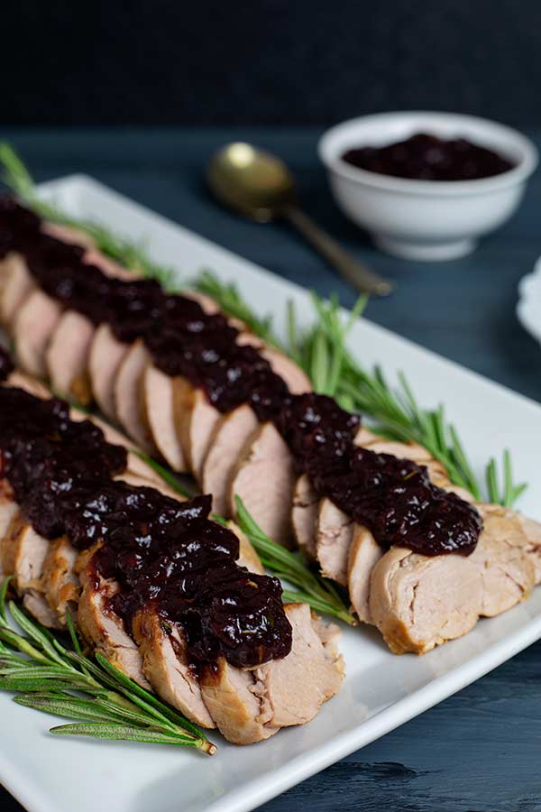 Pork Tenderloin with Cranberry Rosemary Sauce arranged on a white rectangular platter on a dark blue table with dark background
