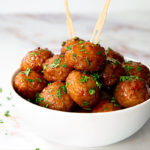 Spicy Sweet & Sour Meatballs in a white bowl sprinkled with parsley on a white marble table