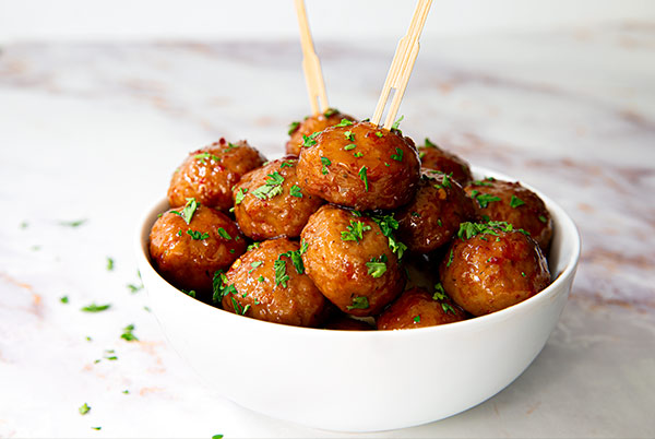 Spicy Sweet & Sour Meatballs in a white bowl sprinkled with parsley on a white marble table