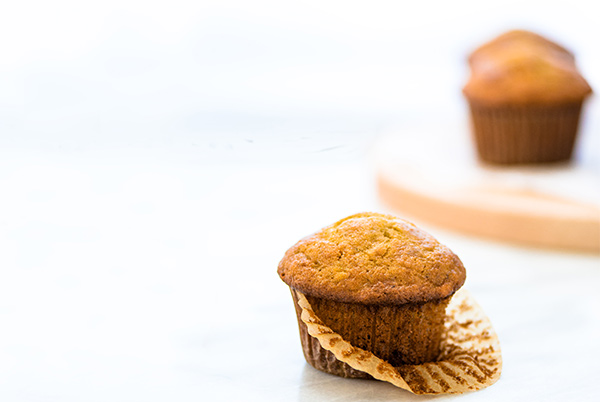 Front view of a Banana Muffin with two banana muffins out of focus to the right on a riser.