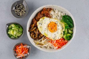 An overhead view of a bowl of Korean Beef Bipimbop in a white bowl with smaller bowls of toppings to the left of it.