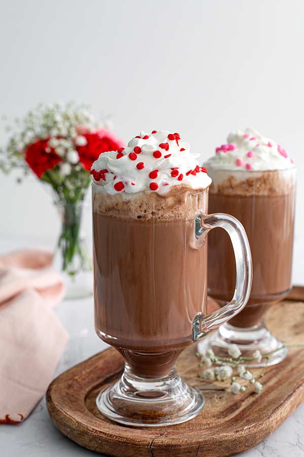 two cups of hot chocolate with whipped cream and red and pink sprinkles on top. 