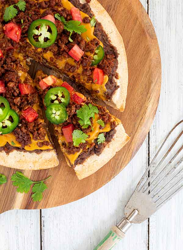 The Plant-based Nacho Pizza with a slice cut out of it on a wooden platter. 